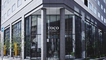 Une entrée d’hôtel moderne, avec des portes en verre, un logo bien visible et une vue dégagée sur la rue.