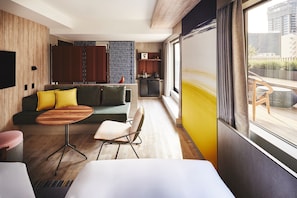 Una habitación de hotel moderna con piso de madera, un sofá con cojines amarillos, una pequeña mesa redonda, una silla, una cama, un área de cocina y un amplio ventanal con vistas a edificios.