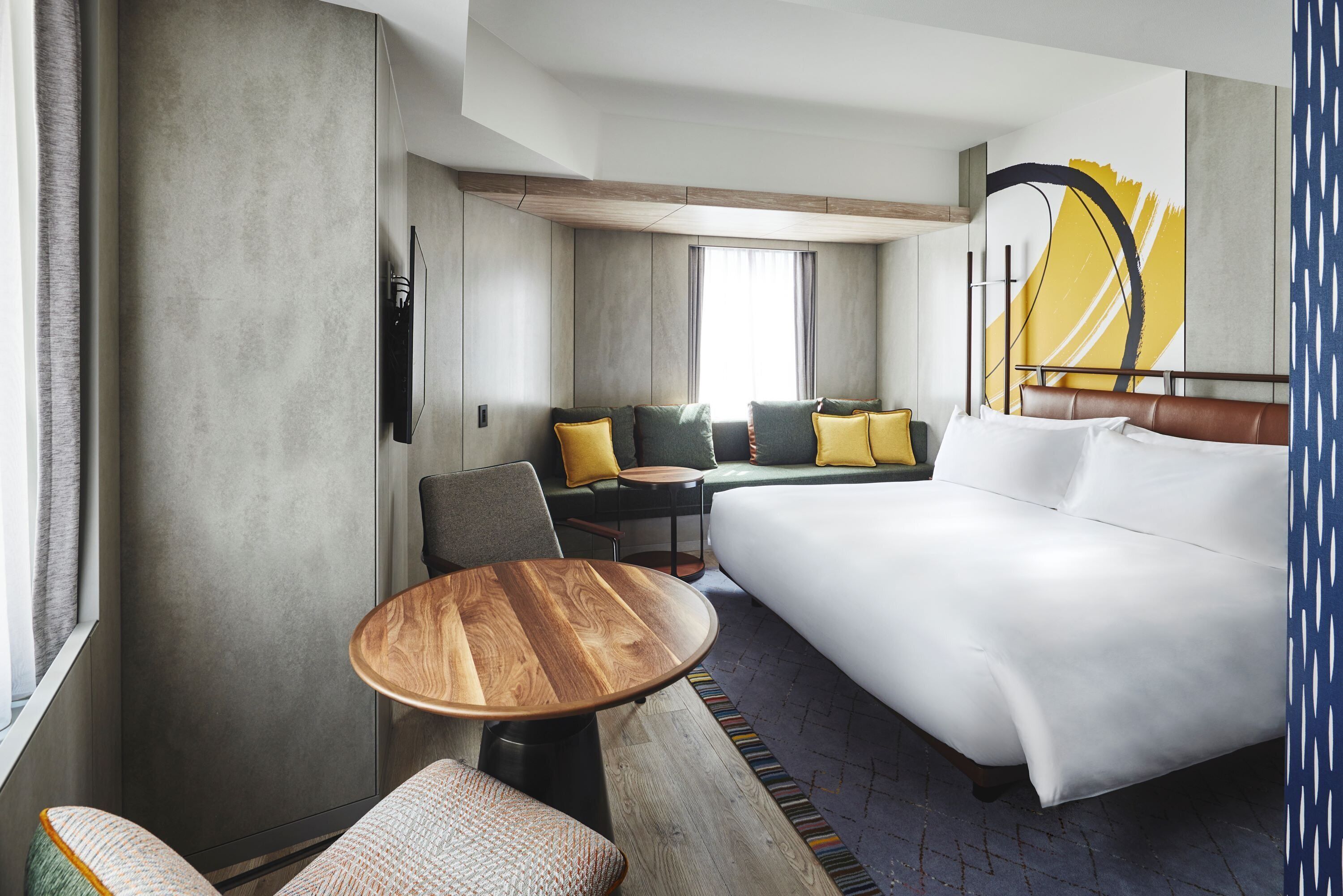 Una habitación de hotel con una cama grande, una mesita, un sofá con cojines, una silla y una ventana con cortinas.