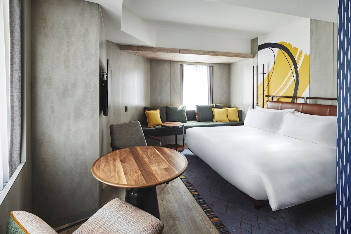 Habitación de hotel con una cama grande, una mesa de madera, un sofá con cojines amarillos y un televisor montado en la pared.