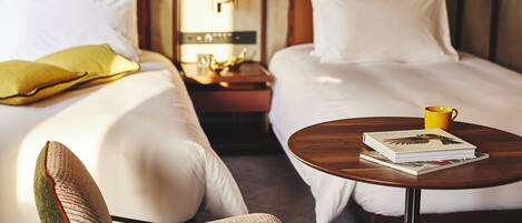 Una habitación de hotel con dos camas, una mesita de noche con una lámpara, una silla y una mesa con un libro y una taza.