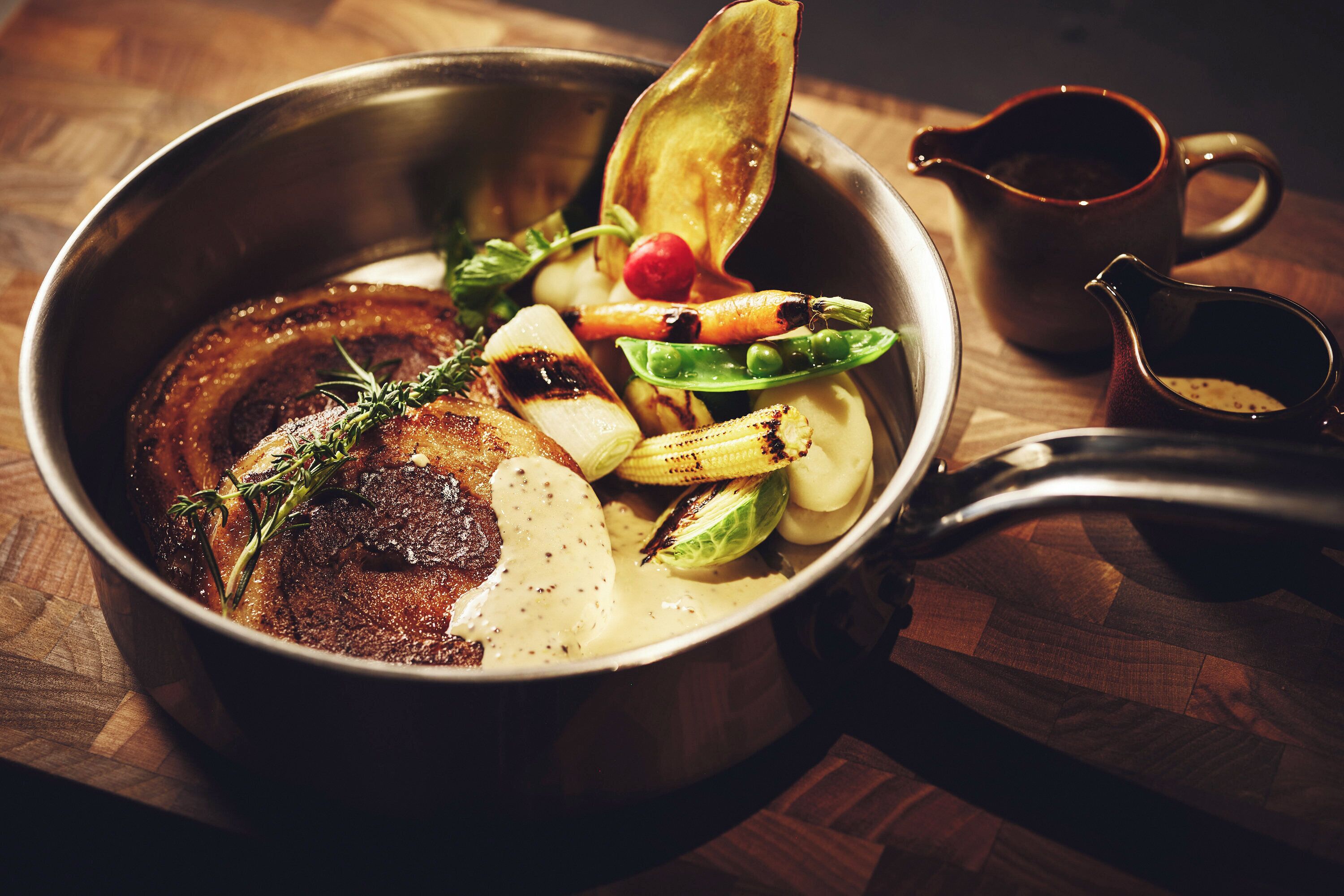 Un plat contenant de la viande rôtie, des légumes et une sauce, accompagné d’un petit pichet et d’une tasse, le tout posé sur une table en bois.