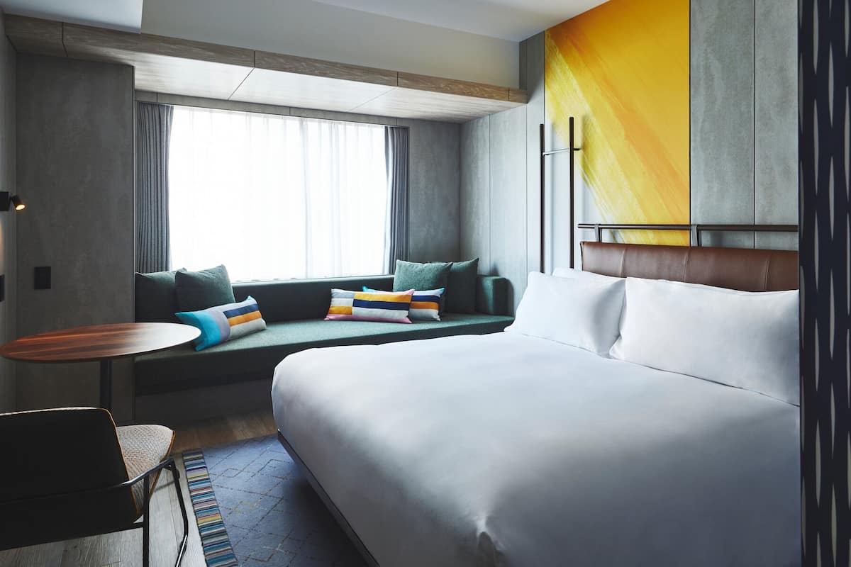 A modern hotel room with a large bed, a sofa with colorful pillows, a small round table, and a view of the outdoors through a window.