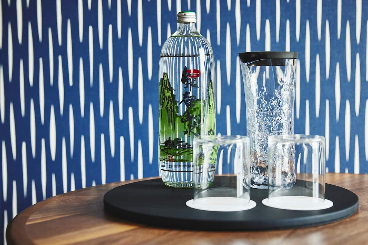 A bottle with a green label and a clear glass container with a black lid on a wooden table against a blue patterned wall.
