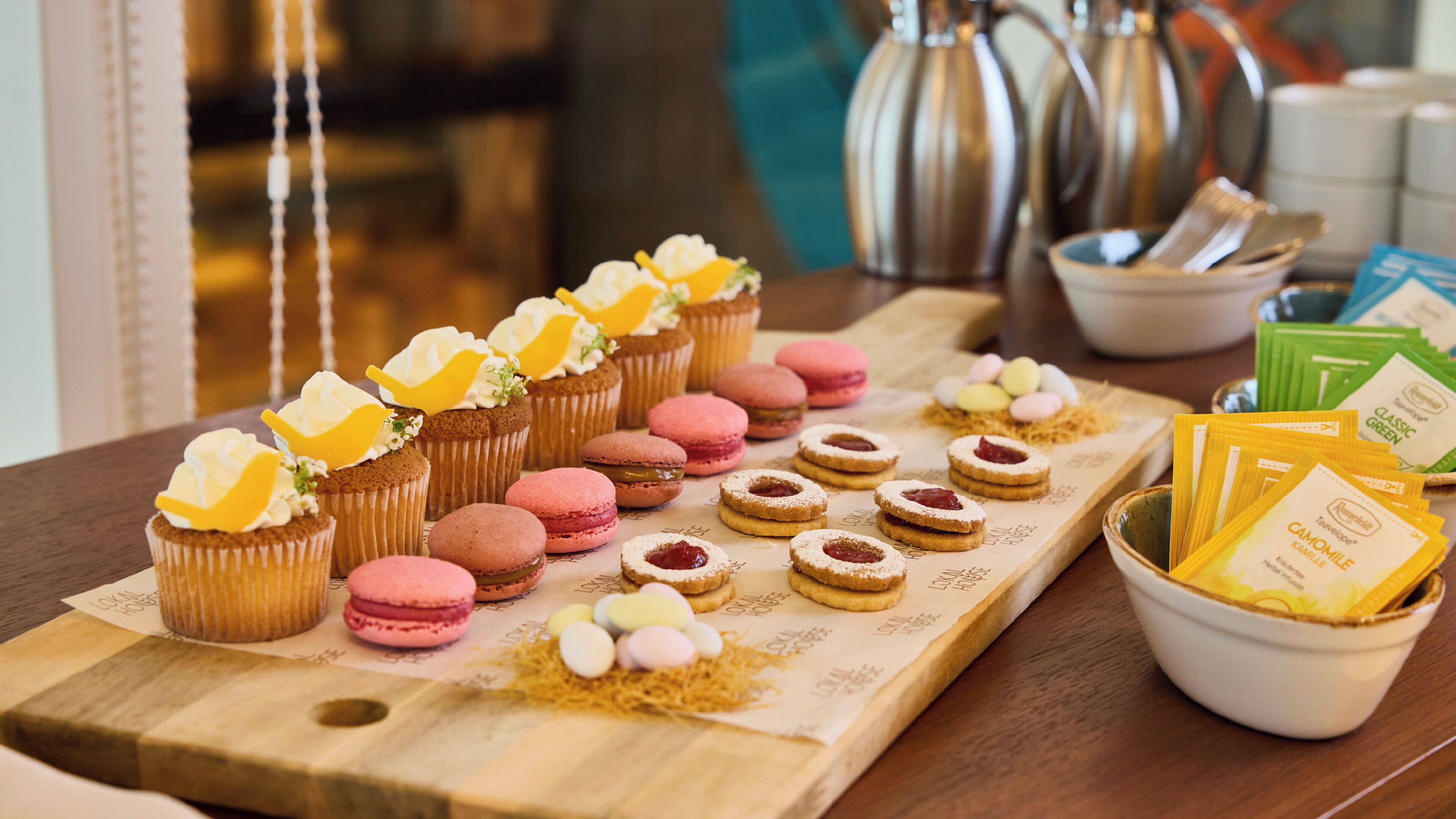 Een houten serveerplank met cupcakes, macarons en koekjes, samen met theezakjes en een schaal thee.