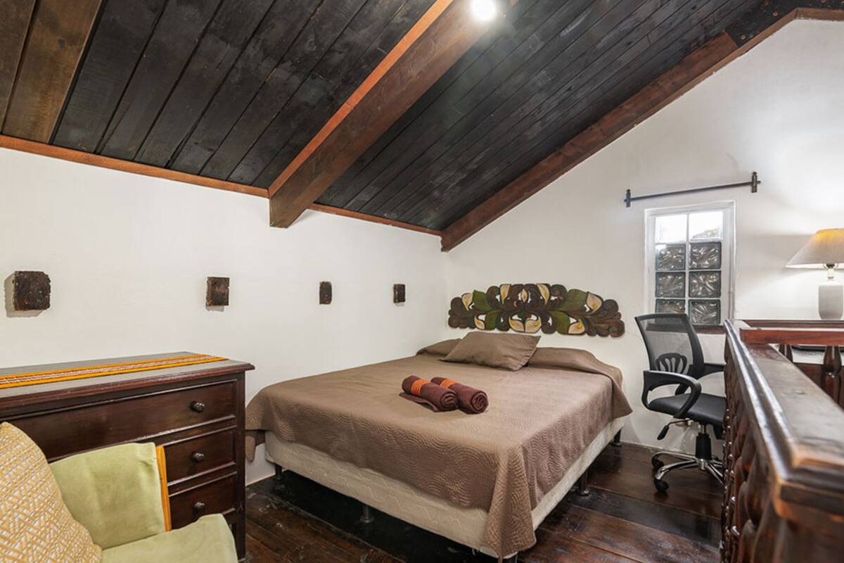 Quarto com teto de madeira, cama com colcha marrom, escrivaninha com luminária e uma cadeira.