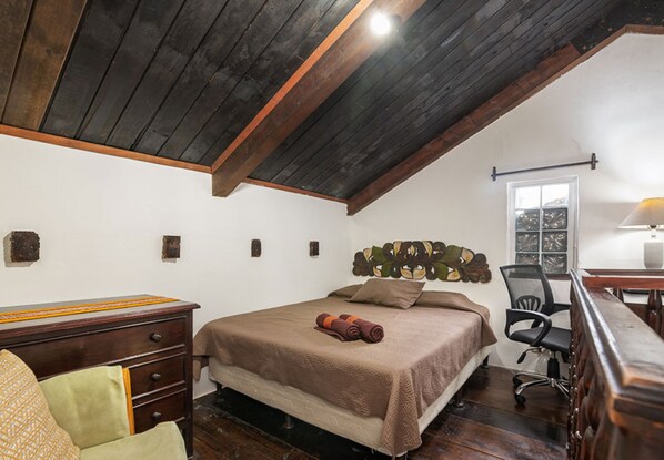 A bedroom with a wooden ceiling, a bed with a brown bedspread, a desk with a lamp, and a chair. - Loft El Chiquito (Antigua Guatemala)