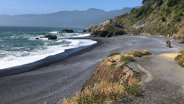 Una carretera costera con una playa a un lado y un promontorio boscoso al otro.