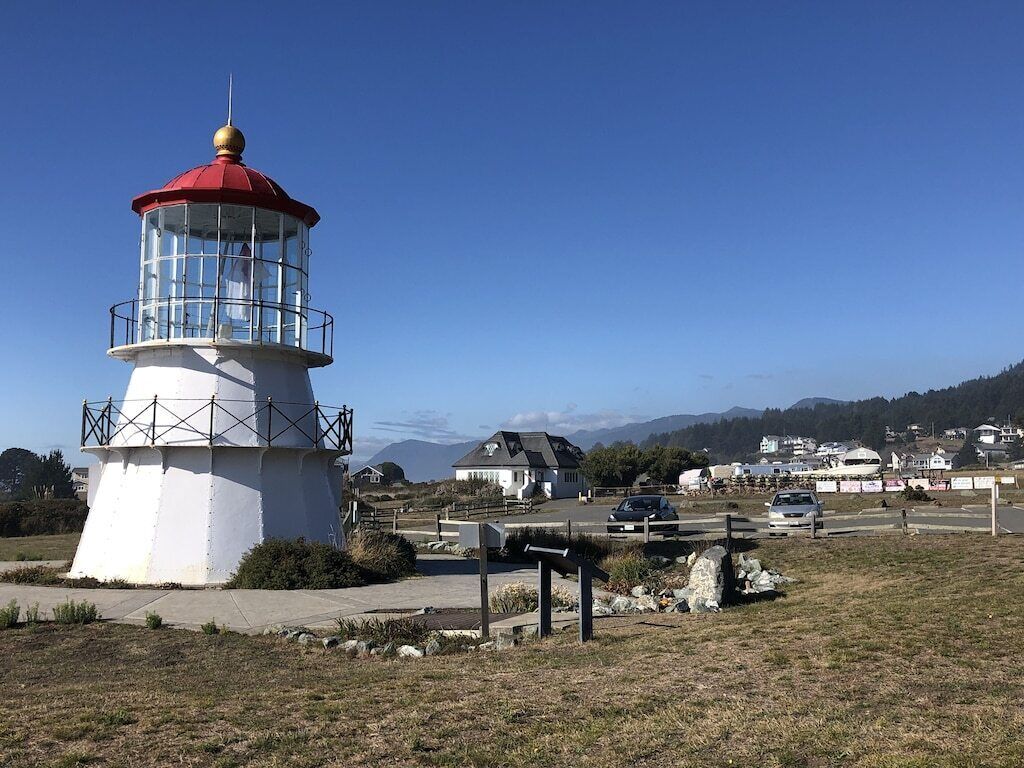 A lighthouse with a red top and white base, surrounded by a grassy area with a parking lot and houses in the background.