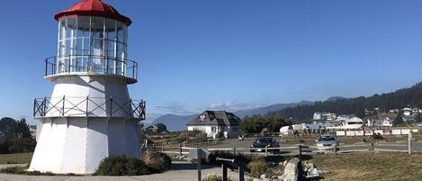 A lighthouse with a red top and white base, surrounded by a grassy area with a parking lot and houses in the background.