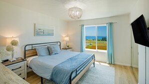 Un dormitorio con cama, mesitas de noche, televisor, ventana con vista al mar y candelabro.