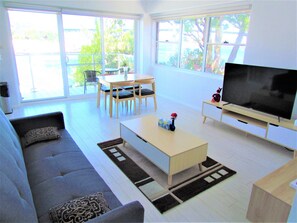 A modern living room with a sofa, coffee table, TV, and a dining area with chairs and a table. - Lumen Harbour – Wake to Sydney’s Brightest Icons (McMahons Point)