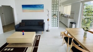 En moderne stue med sofa, sofabord og spisebord. Der er et maleri på væggen, en gulvlampe og udsigt til køkken og balkon.