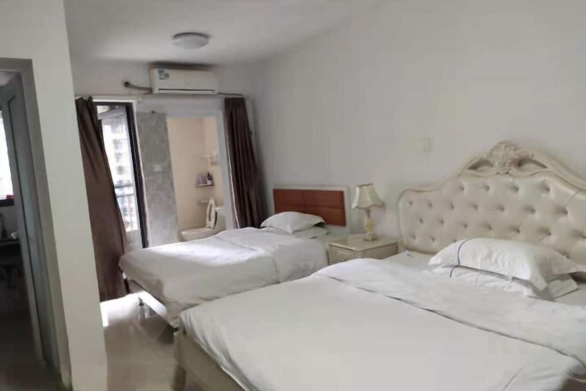 Habitación de hotel con dos camas, un ventanal grande con cortinas, un cabecero blanco y una mesita de noche con una lámpara.