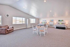 A spacious room with a high ceiling, a dining area with white chairs, a sofa, and a bed with a gray bedspread.