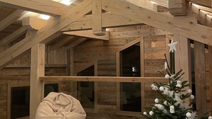 Una habitación cálida en el ático con vigas de madera a la vista, un ventanal grande, un sillón de peluche y un árbol de Navidad decorado.