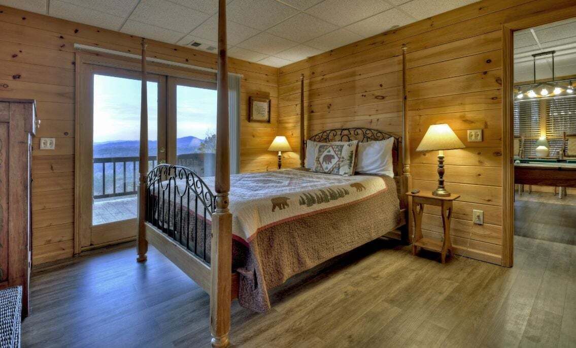 Un dormitorio de cabaña de madera con una cama, dos lámparas de noche, una mesita de noche, un balcón con barandilla y un ventanal grande.