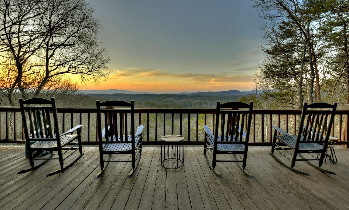 Une terrasse en bois avec trois fauteuils à bascule et une petite table, offrant une vue imprenable sur un paysage pittoresque, avec des arbres et des collines lointaines.