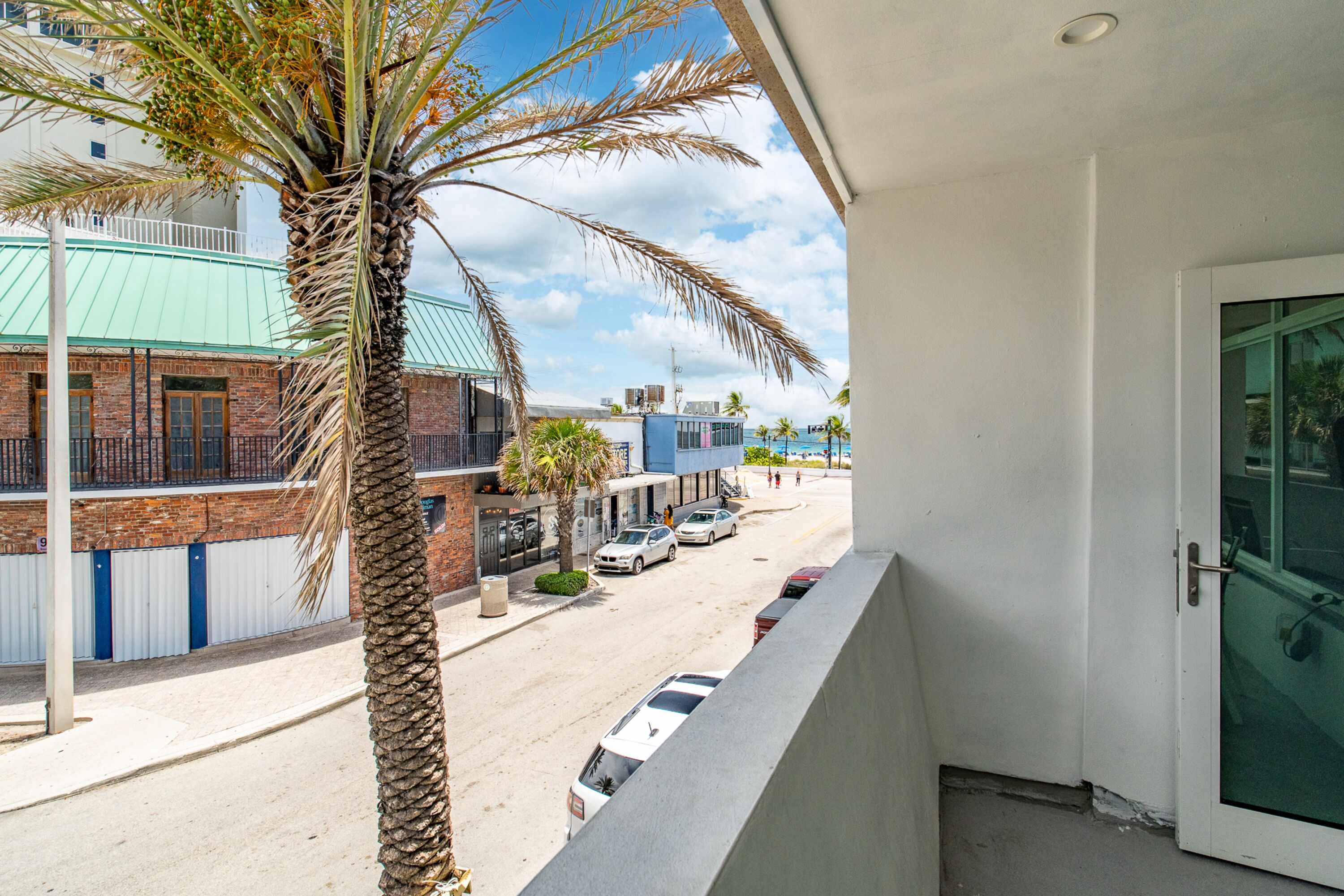A balcony with a view of a street, palm trees, and buildings with a green roof and brick walls.
