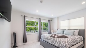 A modern bedroom with a large bed, a flat-screen TV mounted on the wall, and a window with curtains. - Purple Pelican by Opus (Suite 1) (Fort Lauderdale)