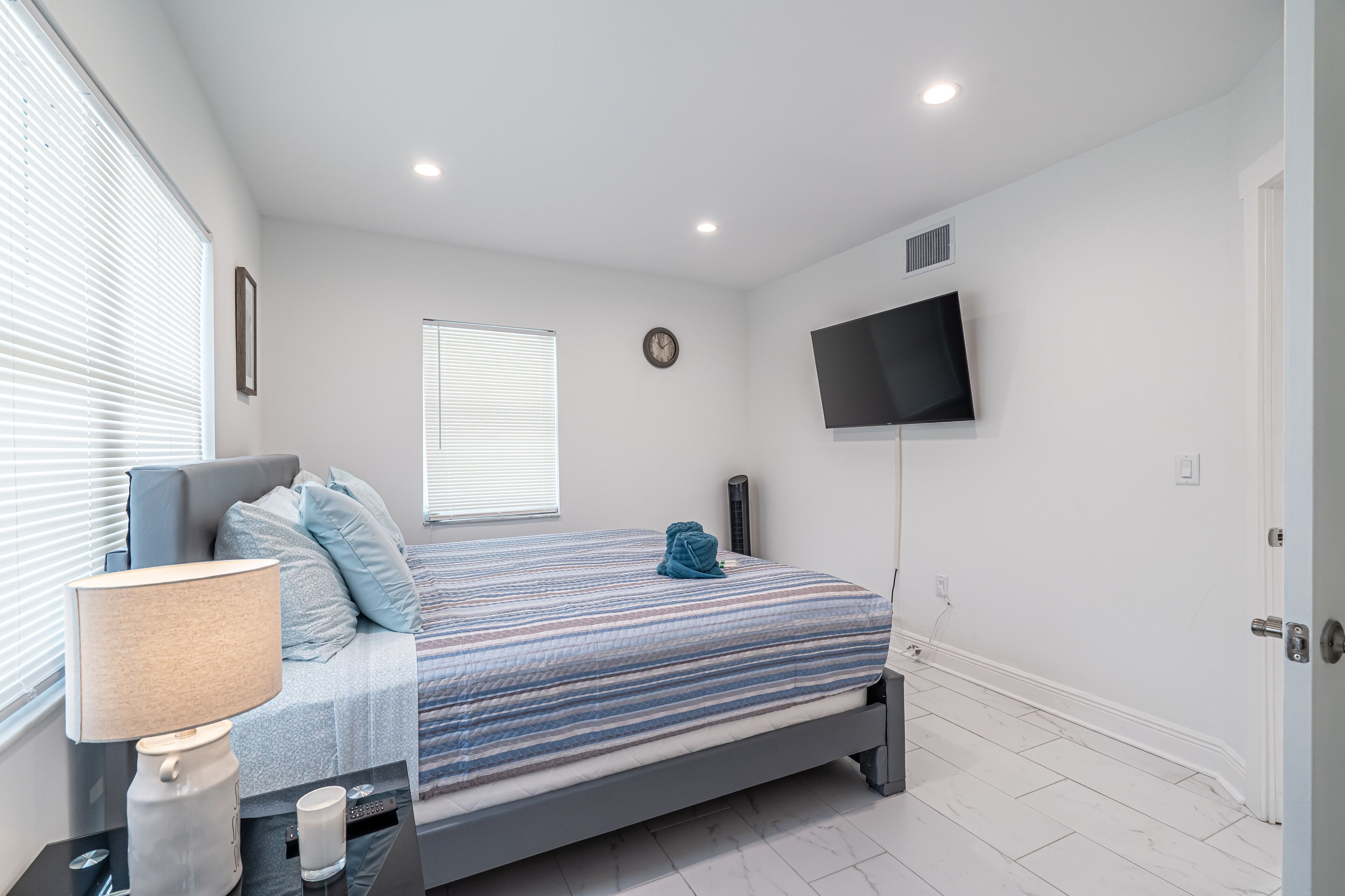 A bedroom with a bed, a TV mounted on the wall, a bedside table with a lamp, and a window with blinds.