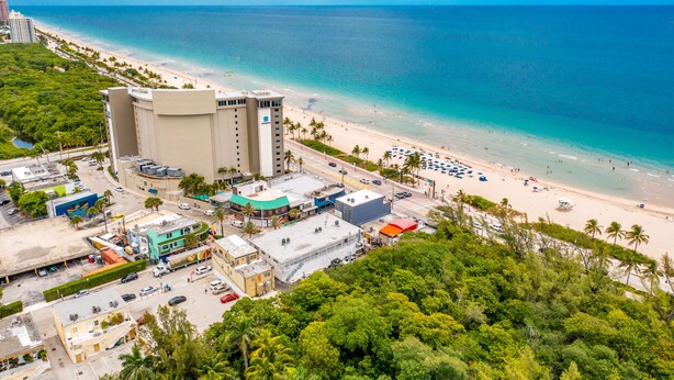 An aerial view of a coastal area with a beach, a hotel with multiple floors, and surrounding greenery. - Purple Pelican by Opus (Suite 1) (Fort Lauderdale)