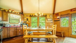 Ein Holzblockhaus-Interieur mit Esstisch, Holzsesseln, einer Kristallleuchtre, und einer Küchenzeile mit Holzschränken und einem Fenster.