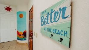 Ein Flur mit einer Tür, einer Wand mit dem Schriftzug 'LIFE IS BETTER AT THE BEACH' und einer Wanddekoration als Sonne.