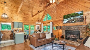 A spacious cabin interior with a stone fireplace, wooden ceiling, and large windows overlooking a forest. - Newly Renovated! NEW deck & SAUNA! 3 King Bedrooms! Hot tub/Arcade. Free tickets (Sevierville)