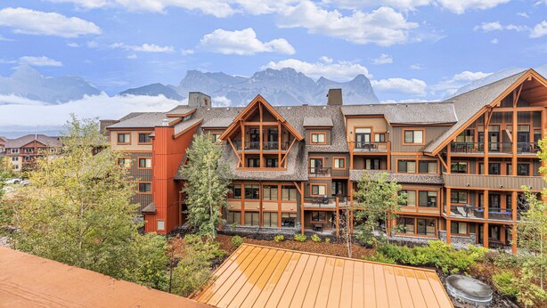 A multi-story lodge with a wooden facade, large windows, and a view of mountains in the background. - Luxury King Suite- Mountain Views/ 2 Balconies/Pool/Hot tub (Canmore)