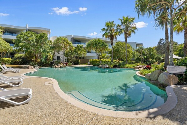 A swimming pool with lounge chairs, surrounded by palm trees and a building in the background.