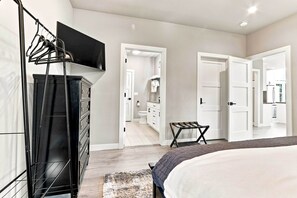 A bedroom with a bed, a TV mounted on the wall, a dresser, and a bathroom visible through an open door.