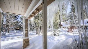 A covered porch with a stone pillar, snow-covered ground, and a view of a house and trees in the background. - Bear Snugglin - Close to Slopes, Hot Tub, Deck, Barbecue (Big Bear Lake)