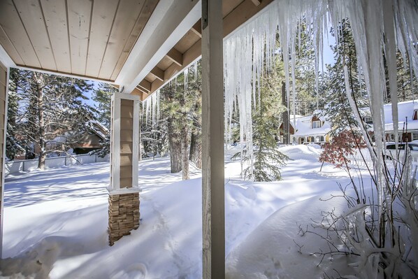 A covered porch with a stone pillar, snow-covered ground, and a view of a house and trees in the background.