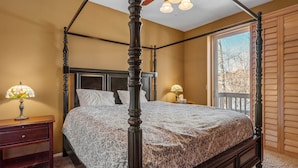 A four-poster bed with a patterned bedspread, a nightstand with a lamp, and a window with a view of trees. - Bear Snugglin - Close to Slopes, Hot Tub, Deck, Barbecue (Big Bear Lake)