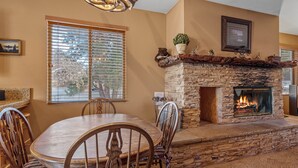 A stone fireplace with a fire, a dining table with chairs, and a window with blinds. - Bear Snugglin - Close to Slopes, Hot Tub, Deck, Barbecue (Big Bear Lake)