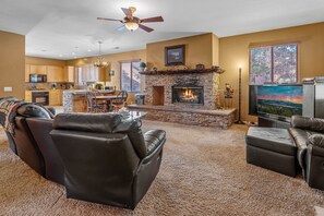 A spacious living room with a fireplace, leather sofas, a television, and a kitchen in the background.