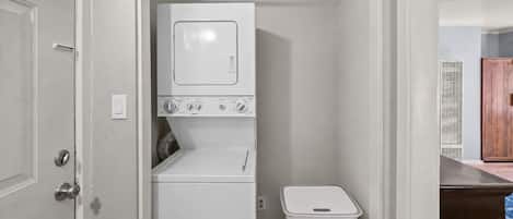 Un lavadero pequeño con lavadora y secadora, un cesto de basura y una puerta que da a otra habitación.