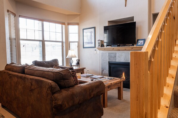 A living room with a brown leather sofa, a wooden coffee table, a fireplace, a TV mounted above it, and a staircase to the right.