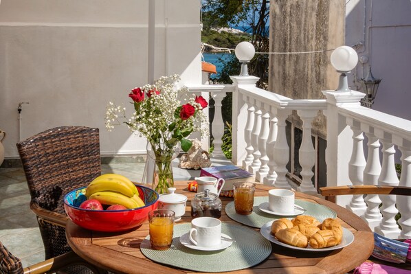 Uma mesa de pequeno-almoço numa varanda, com uma cadeira de vime, frutas, flores e alimentos para o pequeno-almoço.