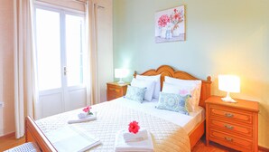 Een netjes opgemaakt bed met een sprei, kussens en een opgerolde handdoek. Er is een nachtkastje met een lamp, een houten hoofdbord en een schilderij aan de muur.