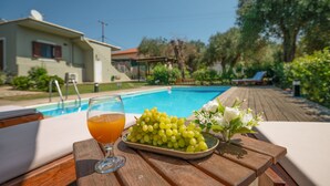 Une table placée au bord de la piscine, avec un verre de jus d’orange, une assiette de raisins verts et un vase de fleurs blanches.
