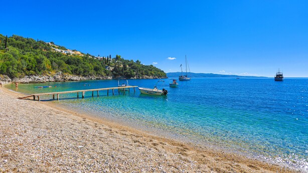 Una playa de guijarros con agua azul cristalina, barcos y un muelle de madera.