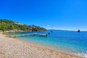 Een kiezelstrand met helderblauw water, boten en een houten pier.