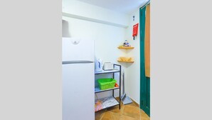 Una cocina compacta con un refrigerador blanco, una pequeña encimera con hervidor y tostadora, una cesta verde en un estante y un aviso rojo de seguridad contra incendios en la pared.