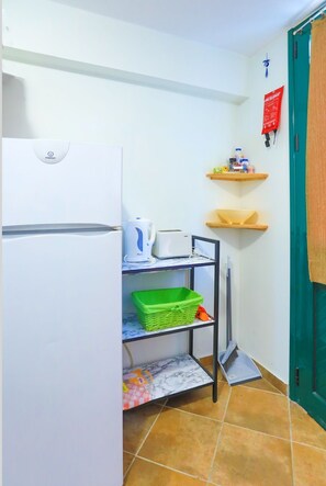 A compact kitchen with a white refrigerator, a small countertop with a kettle and toaster, a green basket on a shelf, and a red fire safety notice on the wall.