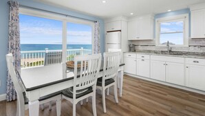 Une cuisine avec des meubles blancs, une table à manger avec des chaises, une vue sur la mer et un balcon avec une balustrade.