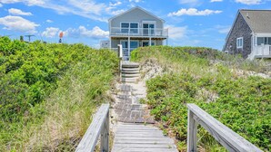 Une maison située sur la côte, dotée d’un escalier en bois menant à un balcon, le tout entouré d’une végétation luxuriante.