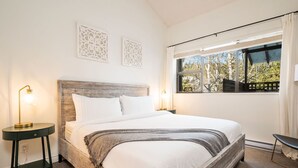 Ein Schlafzimmer mit einem hölzernen Kopfteil, einem großen Bett mit weißer Bettwäsche, zwei Wandpaneelen zur Dekoration, einem Nachttisch mit Lampe sowie einem Fenster mit Blick auf Bäume und ein Gebäude.