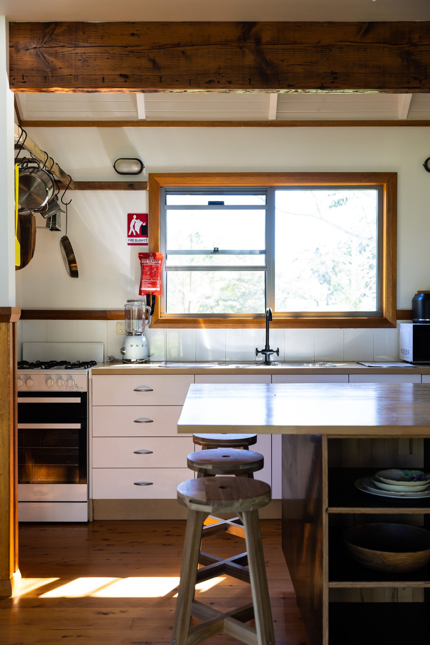 A kitchen with a wooden ceiling, a window, a stove, a sink, and a stool.